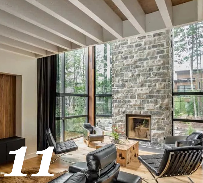 Burası yüksek tavanlı ve cam duvarlarla çevrili taş şömineli oturma odası, ahşap tavanı duvar renginde kirişler tamamlıyor, aydınlatma tamamen doğal.